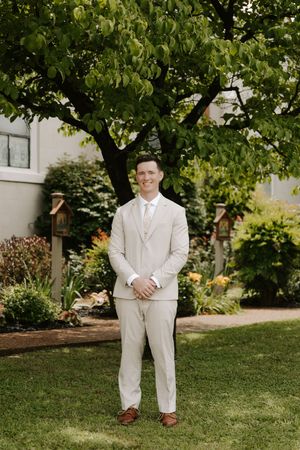 Groom in beige suit smiling outdoors in lush garden setting.