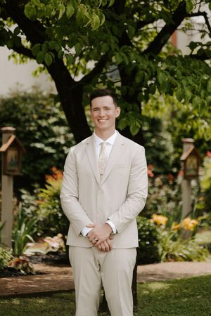 Smiling groom in beige suit with tie stands in lush garden with hands clasped.