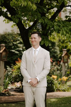 Groom in tan suit smiles softly while standing in colorful garden beneath large tree.