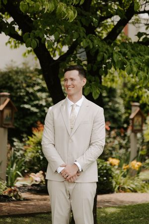 Smiling groom in beige suit standing in lush garden before wedding ceremony.