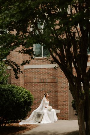 Bride in white gown and veil sits gracefully on a bench outside a brick building surrounded by lush greenery.