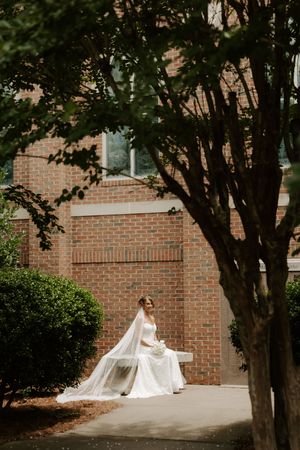 Bride in a white gown and long veil sits on a bench outside a brick building surrounded by lush greenery.