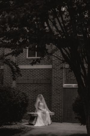 Bride in white gown and veil sitting alone on bench outside brick building in black and white photo.