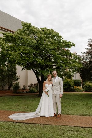 Bride and groom posing together on green lawn with white wedding dress and beige suit.