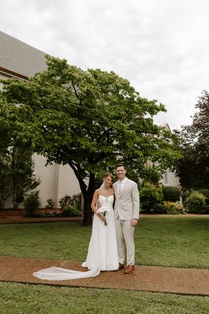 Bride and groom pose together on a garden path in front of a large tree on their wedding day.