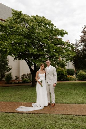 Bride and groom pose together in a lush garden, she in a white gown with train, he in a beige suit.