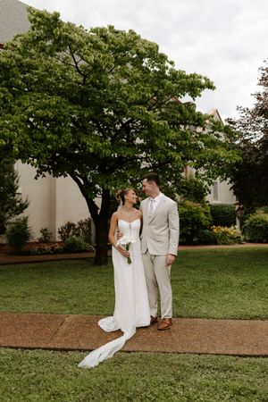 Bride and groom stand together on a garden path, smiling at each other, with a lush green tree backdrop.