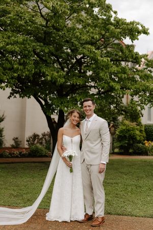 Bride in white strapless gown with long veil and groom in tan suit pose outdoors on green lawn near large tree.