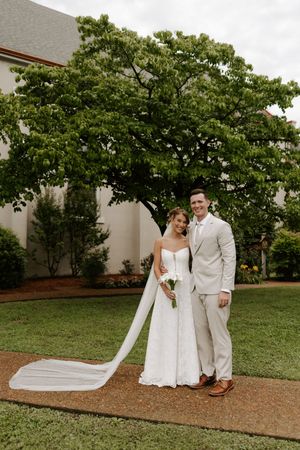 Bride and groom pose together on garden path, her cathedral veil trailing behind, both smiling in elegant wedding attire.