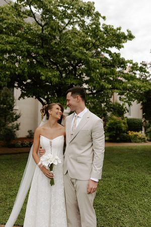 Bride and groom share a joyful moment on a green lawn, she holds white calla lilies in her lace wedding gown.