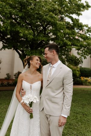 Smiling bride and groom share a tender moment outdoors, she holds white calla lilies, wearing a strapless gown and veil.