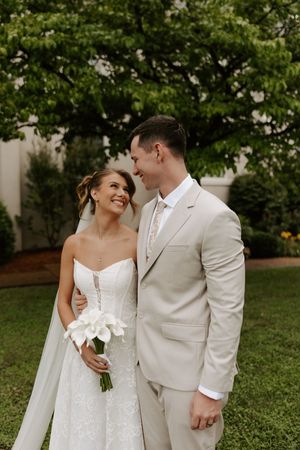 Smiling bride holding white calla lily bouquet leans into groom in tan suit outdoors on their wedding day.