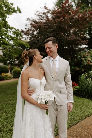 Smiling newlyweds share a tender moment outdoors, bride holding white calla lily bouquet in lace gown with veil.