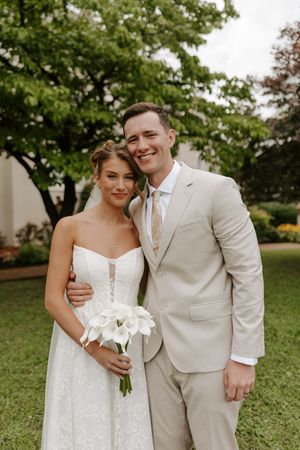 Smiling bride holding white calla lily bouquet and groom in beige suit embrace outdoors in lush garden setting.