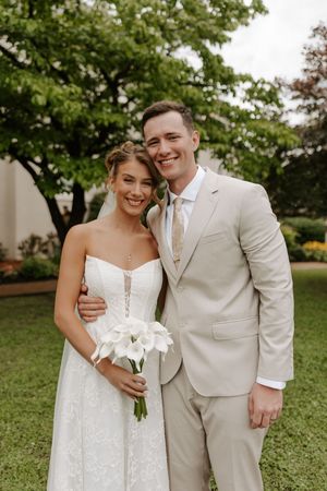 Happy bride in strapless lace gown holds calla lily bouquet beside groom in tan suit, posing in lush garden setting.