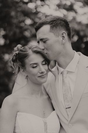 Black and white close-up of groom kissing bride on temple, bride smiling softly in strapless wedding gown.