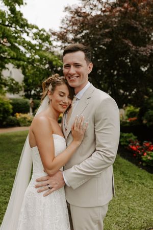 Groom embraces bride from behind as she rests her hand on his chest, both smiling in a garden setting.