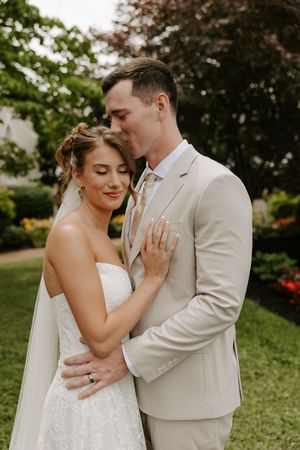 Groom in tan suit kisses bride's temple as she smiles with eyes closed in lace gown and veil.