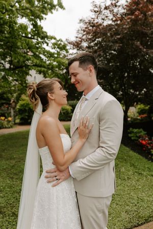 Bride and groom share a romantic moment in a lush garden, bride in strapless lace gown with veil, groom in beige suit.