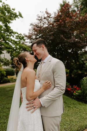 Smiling bride and groom in tan suit nearly kiss while embracing in lush garden setting.