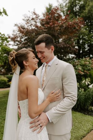Smiling bride and groom embracing in garden, groom in tan suit with floral tie, bride in strapless lace gown.