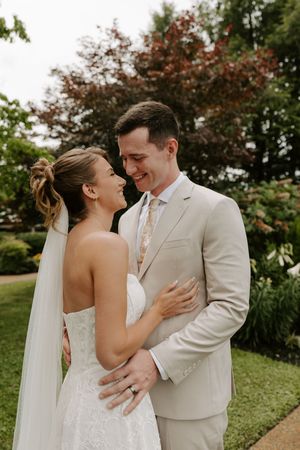 Bride in strapless lace gown and groom in beige suit share a joyful smile outdoors.