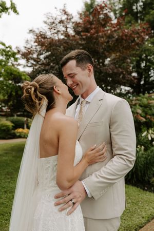 Smiling groom in tan suit embraces bride in strapless lace wedding dress with veil in a green garden setting.