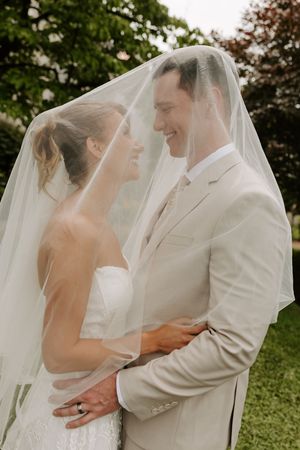 Smiling bride and groom share intimate moment together under flowing white bridal veil in garden setting.
