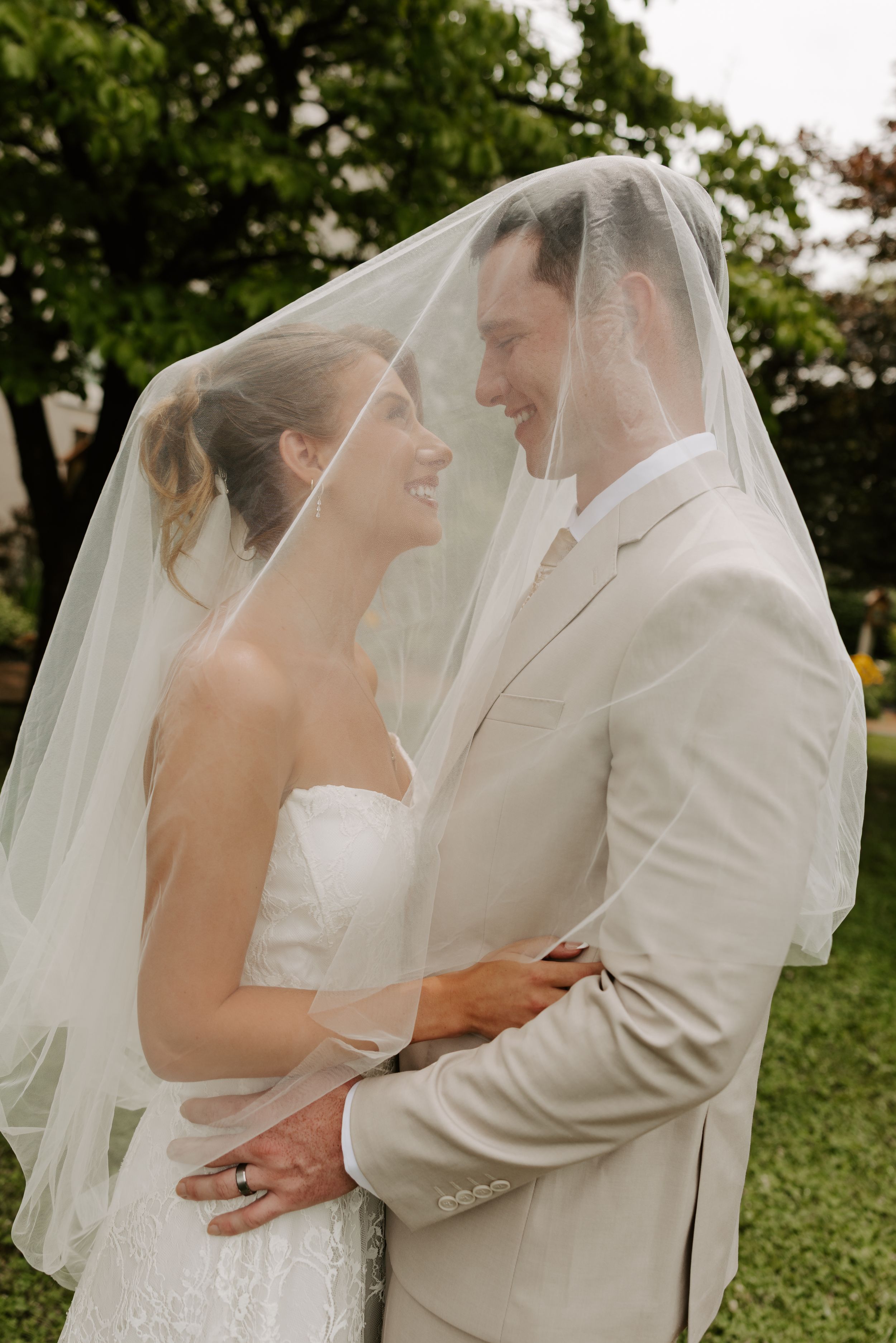 Smiling bride and groom stand close together draped under a sheer bridal veil outdoors on their wedding day.