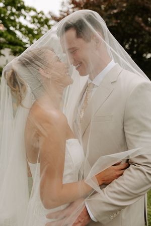 Bride and groom share romantic moment under wedding veil, smiling at each other outdoors.