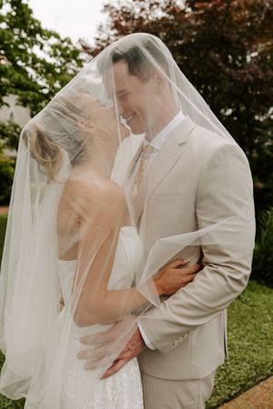 Romantic bride and groom share a close moment under a flowing bridal veil outdoors.