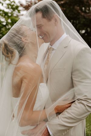 Smiling bride and groom lean in for a kiss beneath a sheer bridal veil in a garden.