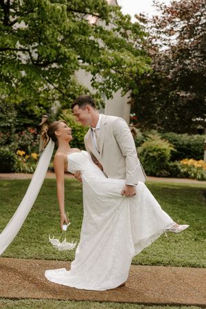 Groom in tan suit lifts laughing bride in white lace gown on garden pathway outdoors.