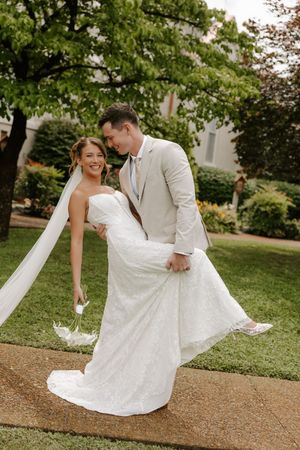 Laughing groom in tan suit lifts bride's lace gown train as couple walks along garden pathway together.