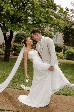 Groom dips smiling bride in white lace wedding gown with flowing veil in garden setting.