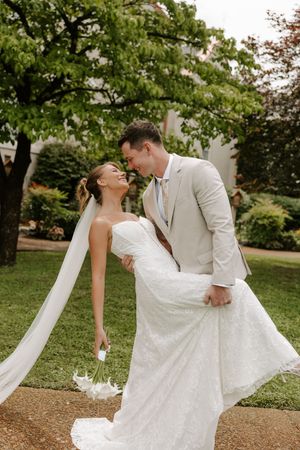 Groom lifts bride as they laugh together in a lush garden, her long veil flowing in the breeze.
