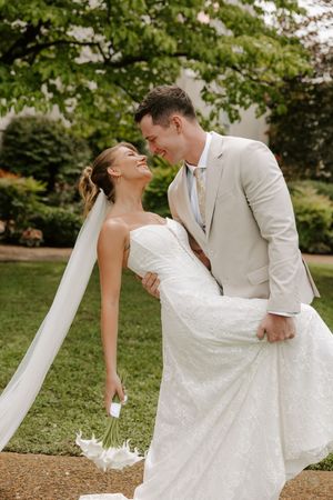 Groom in beige suit dips smiling bride in white lace gown and veil outdoors.