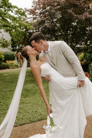 Groom dips bride for a kiss outdoors, bride holding white calla lily bouquet, wearing lace gown.