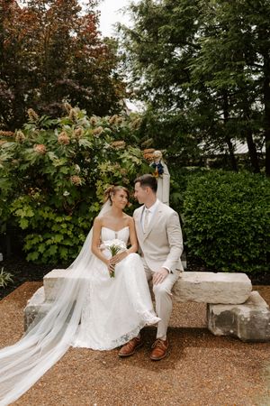 Bride and groom sit on stone bench in garden, bride holding bouquet in lace gown with flowing veil, groom in beige suit.