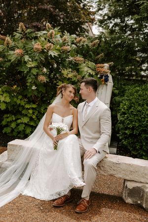 Bride and groom sit on stone bench smiling at each other surrounded by lush greenery.