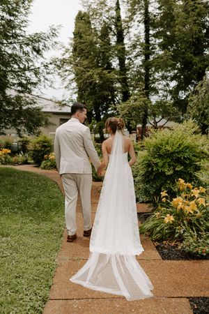 Bride and groom walk hand in hand through lush garden path, bride wearing flowing white gown with veil.