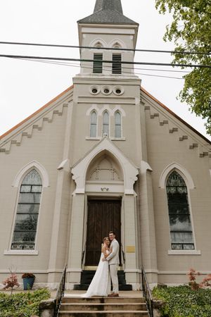 Bride and groom pose on steps of elegant white Gothic Revival church with steeple on their wedding day.