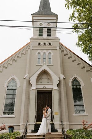 Bride and groom share a romantic kiss on the steps of a white Gothic-style church with a tall steeple.