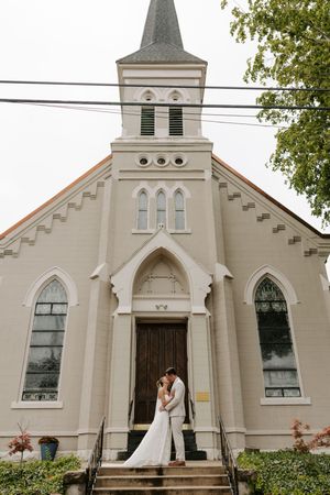 Bride and groom kissing on church steps in front of a white Gothic-style church with a steeple on their wedding day.