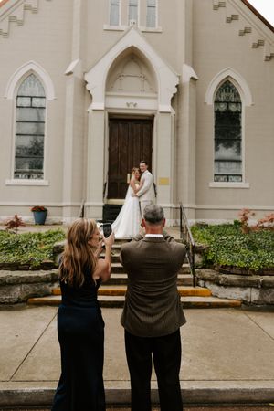 Guests photograph bride and groom posing on steps of white Gothic church entrance doors.