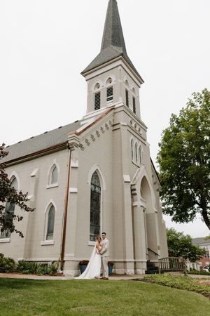 Bride and groom embrace in front of a classic white church with tall steeple on their wedding day.