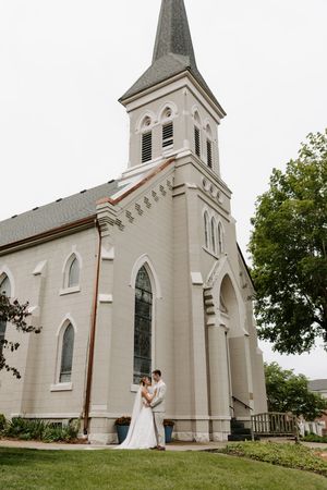 Bride and groom pose romantically in front of elegant white church with tall steeple.