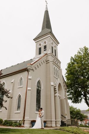 Bride and groom embrace outside a white historic church with a tall steeple on their wedding day.