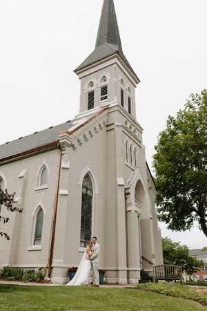 Bride and groom pose together on the lawn beside a grand white church with a tall steeple on their wedding day.