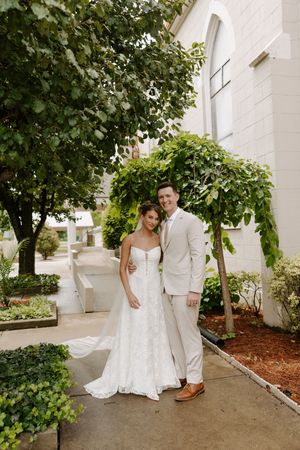 Smiling bride and groom pose together outside white church surrounded by lush green trees.